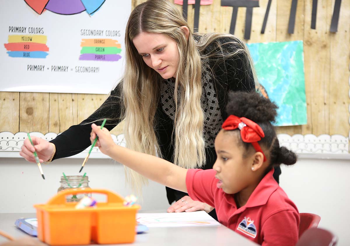 HSA Teacher smiles while kneeling beside a young student in a classroom setting.